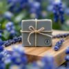 alt="Handcrafted slate gray sea clay soap with tea tree and lavender, wrapped in natural twine on wooden butcher block, accented with dried lavender sprigs against soft-focused Texas bluebonnets"