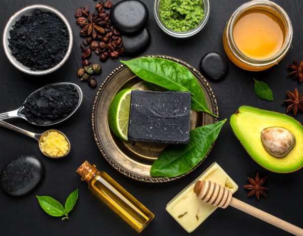 Natural charcoal soap bar on vintage brass plate with ingredients including activated charcoal, lime, avocado, essential oils, coffee beans, and honey on dark slate surface